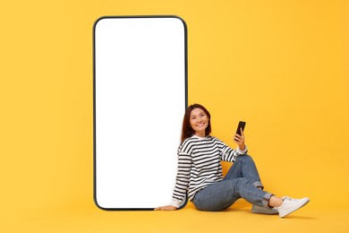 Happy smiling woman with smartphone sitting near big mobile phone with blank screen on golden background. Mockup for design