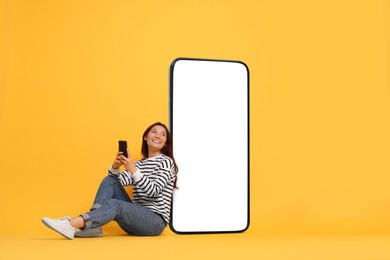 Image of Happy smiling woman with smartphone sitting near big mobile phone with blank screen on golden background. Mockup for design