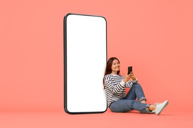 Image of Happy smiling woman using smartphone while sitting near big mobile phone with blank screen on coral background. Mockup for design