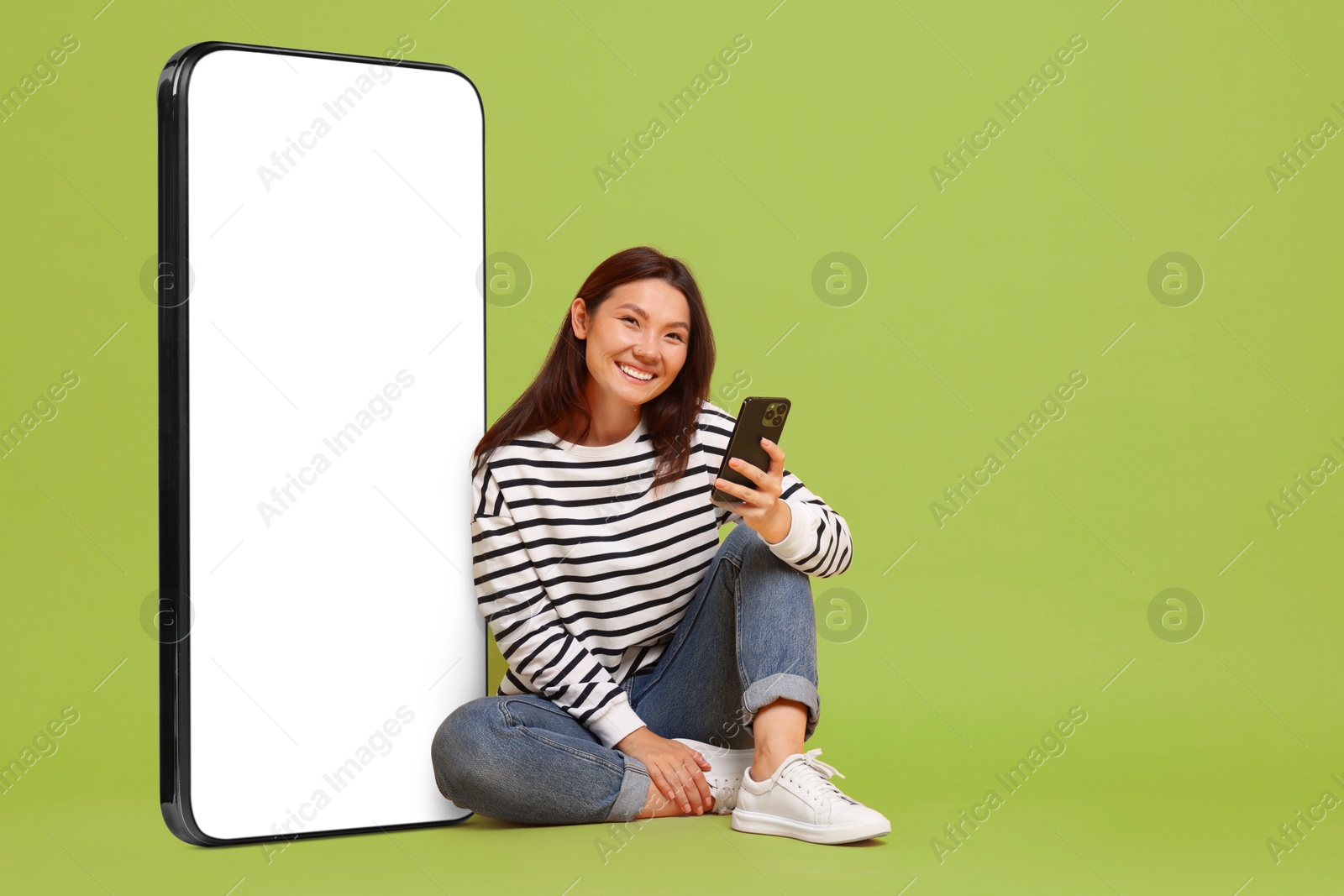 Image of Happy smiling woman with smartphone sitting near big mobile phone with blank screen on light green background. Space for design