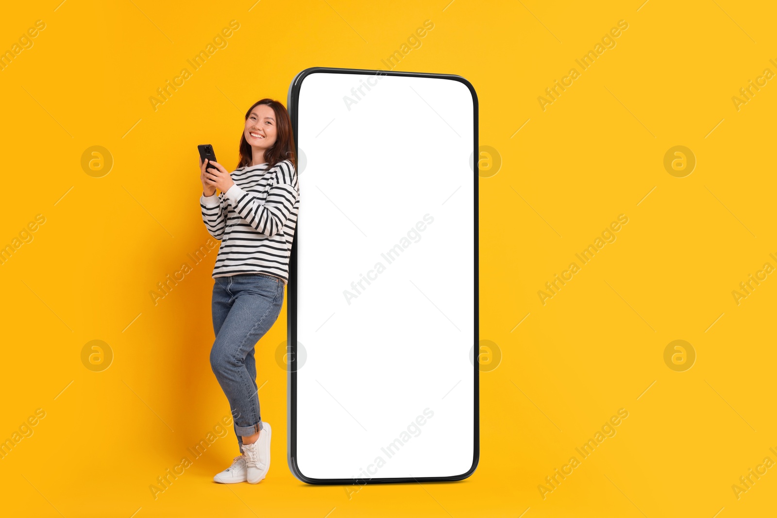 Image of Happy smiling woman with smartphone standing near big mobile phone with blank screen on orange background. Mockup for design