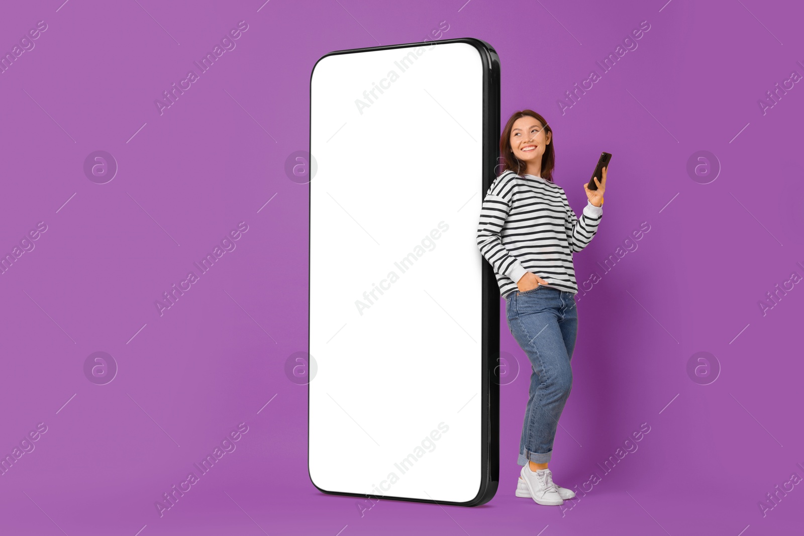 Image of Happy smiling woman with smartphone standing near big mobile phone with blank screen on violet background. Mockup for design