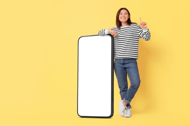 Happy smiling woman and big mobile phone with blank screen on pale yellow background. Mockup for design