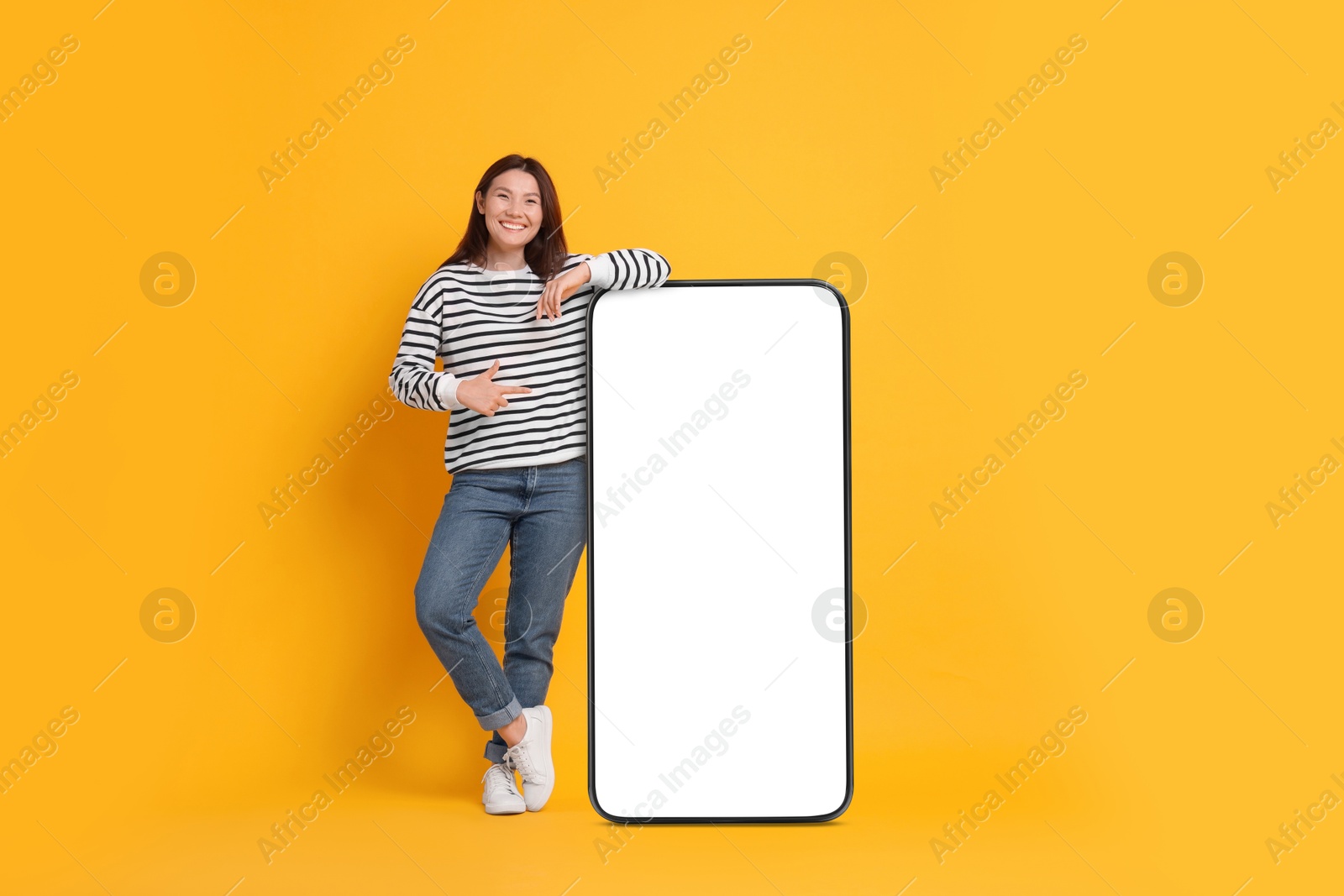 Image of Happy smiling woman pointing at big mobile phone with blank screen on orange background. Mockup for design