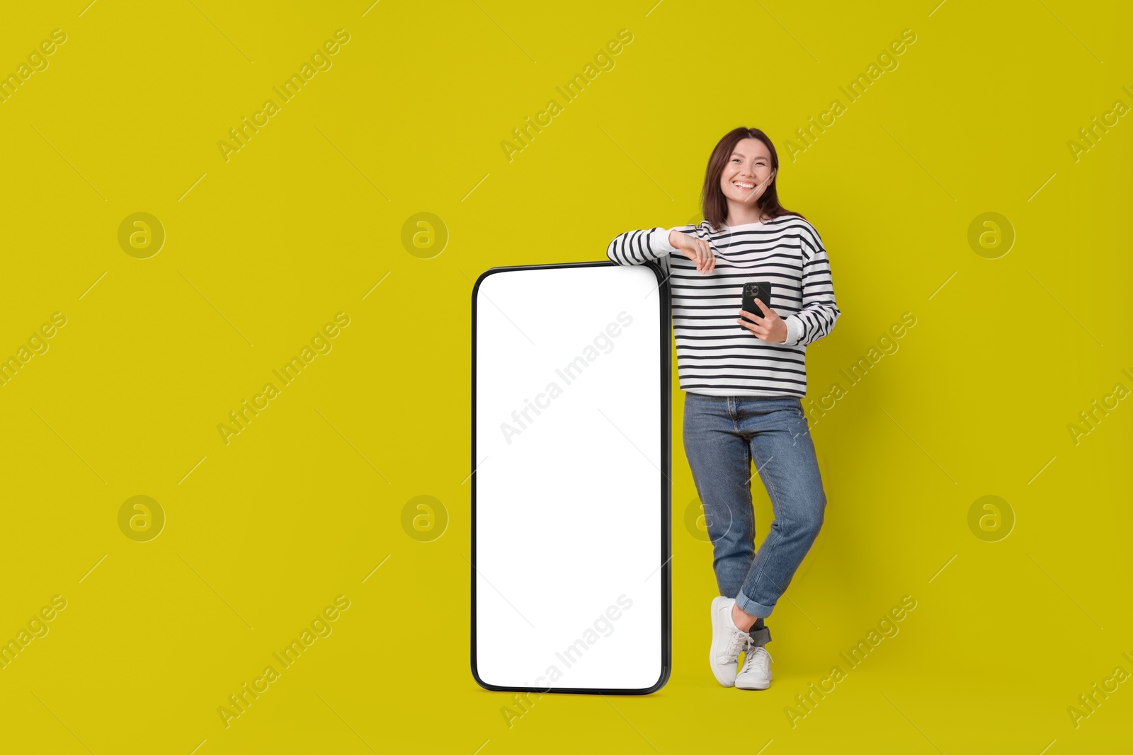 Image of Happy smiling woman with smartphone standing near big mobile phone with blank screen on color background. Mockup for design