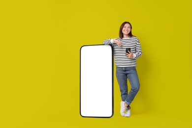 Image of Happy smiling woman with smartphone standing near big mobile phone with blank screen on color background. Mockup for design