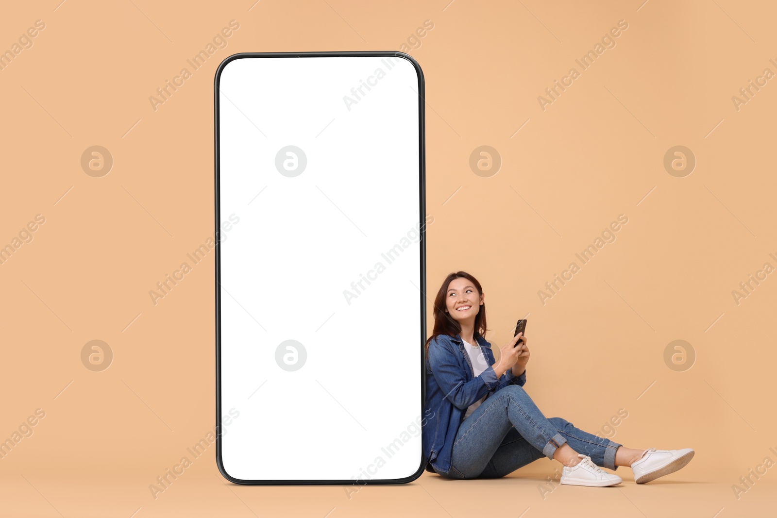 Image of Happy smiling woman with smartphone sitting near big mobile phone with blank screen on dark beige background. Mockup for design