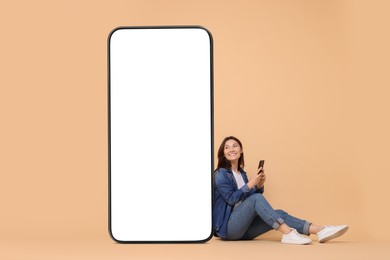 Image of Happy smiling woman with smartphone sitting near big mobile phone with blank screen on dark beige background. Mockup for design