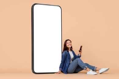 Happy smiling woman with smartphone sitting near big mobile phone with blank screen on dark beige background. Mockup for design