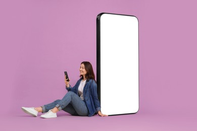 Image of Happy smiling woman using smartphone while sitting near big mobile phone with blank screen on lilac background. Mockup for design