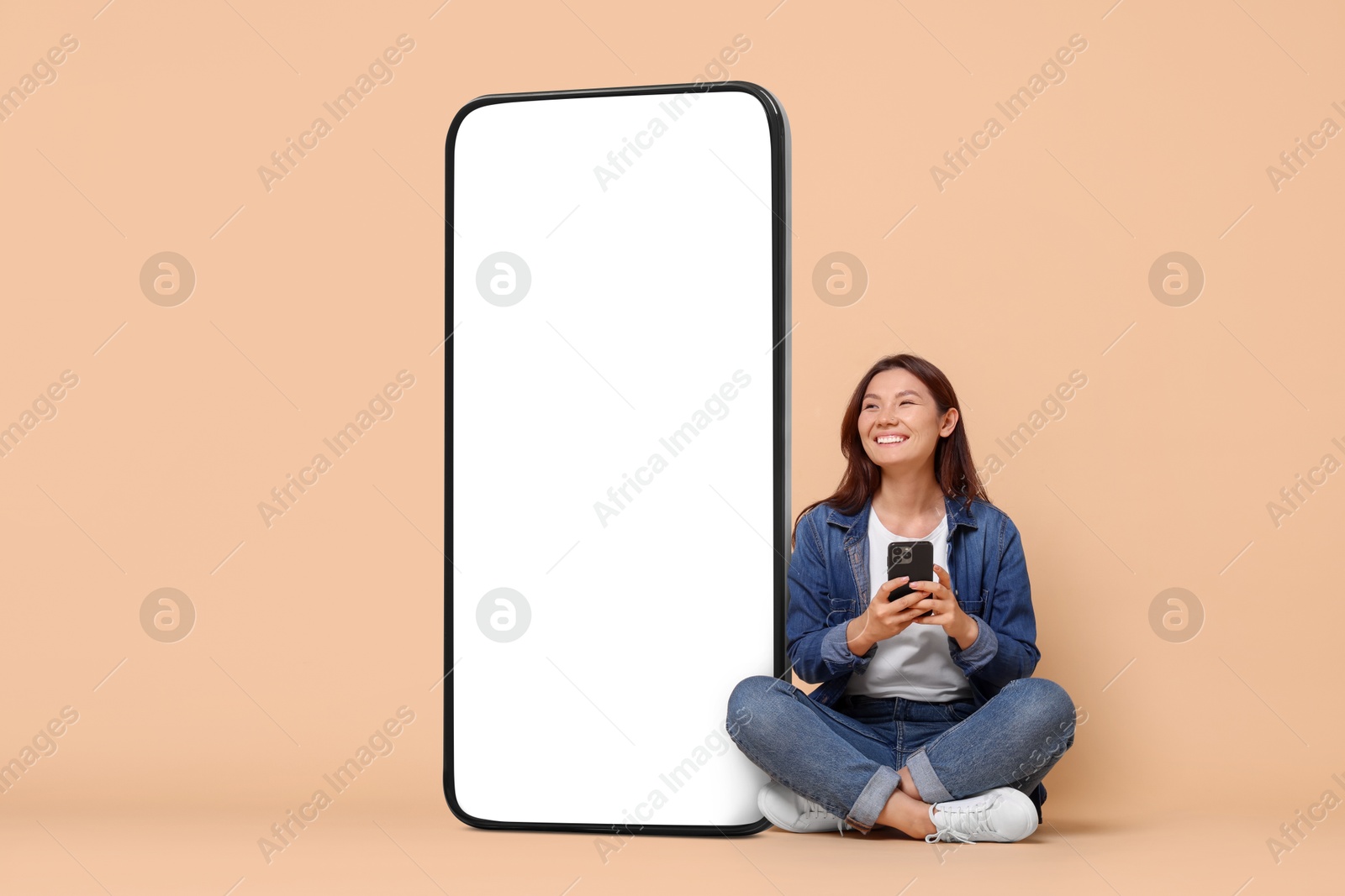 Image of Happy smiling woman with smartphone sitting near big mobile phone with blank screen on dark beige background. Mockup for design