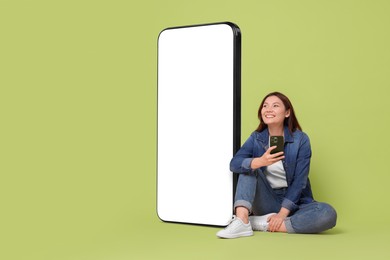 Happy smiling woman with smartphone sitting near big mobile phone with blank screen on light green background. Space for design