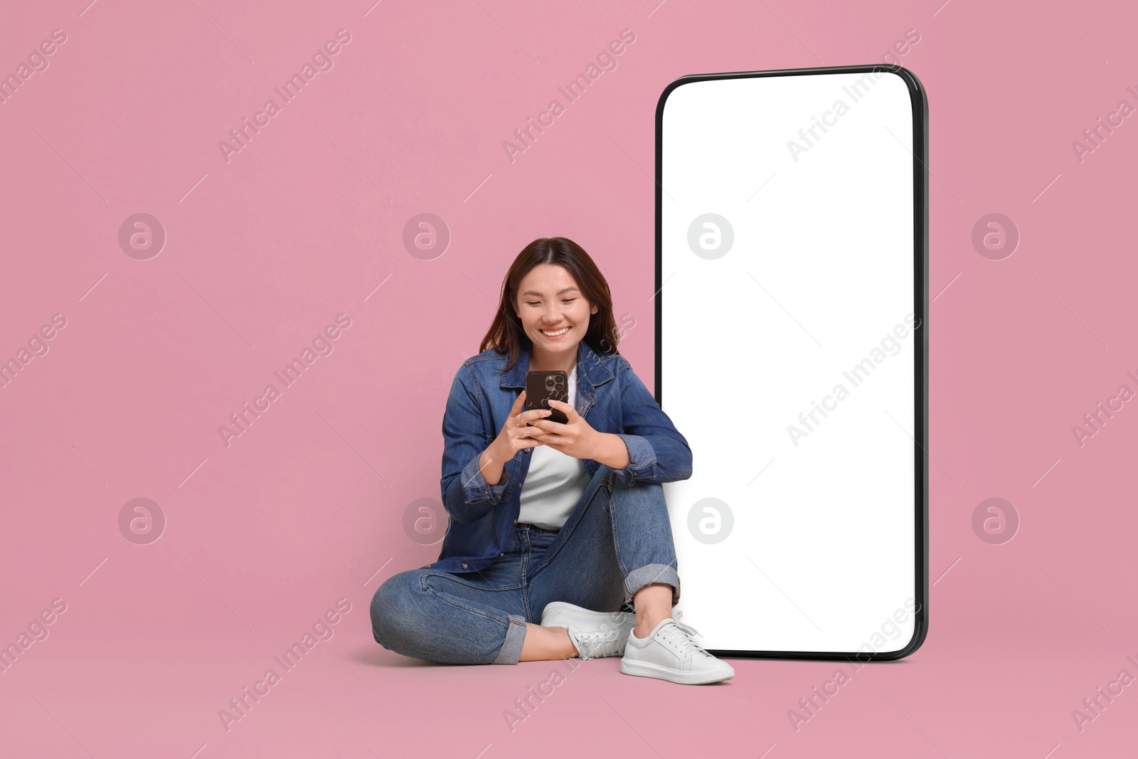 Image of Happy smiling woman using smartphone while sitting near big mobile phone with blank screen on dusty rose background. Mockup for design