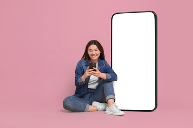 Image of Happy smiling woman using smartphone while sitting near big mobile phone with blank screen on dusty rose background. Mockup for design