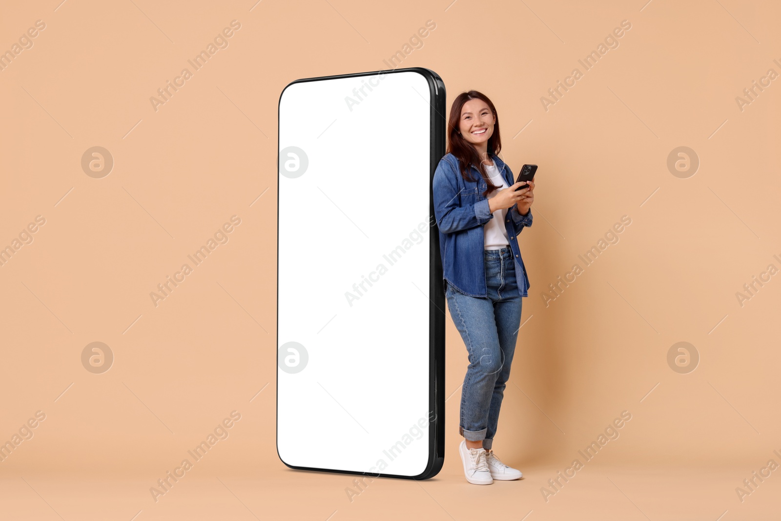 Image of Happy smiling woman with smartphone standing near big mobile phone with blank screen on dark beige background. Mockup for design