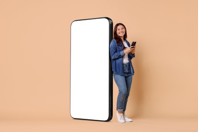 Image of Happy smiling woman with smartphone standing near big mobile phone with blank screen on dark beige background. Mockup for design