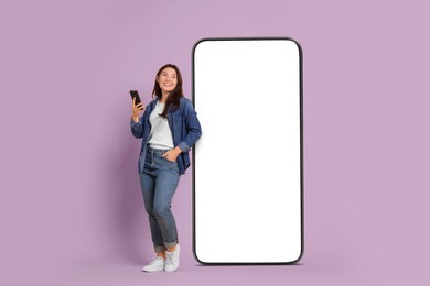 Happy smiling woman with smartphone standing near big mobile phone with blank screen on lilac background. Mockup for design