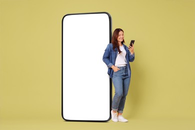 Happy smiling woman using smartphone while standing near big mobile phone with blank screen on color background. Mockup for design