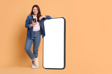 Image of Happy smiling woman with smartphone standing near big mobile phone with blank screen on dark beige background. Mockup for design