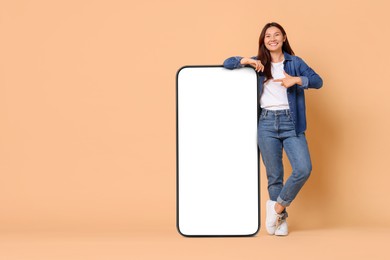 Image of Happy smiling woman pointing at big mobile phone with blank screen on dark beige background. Mockup for design