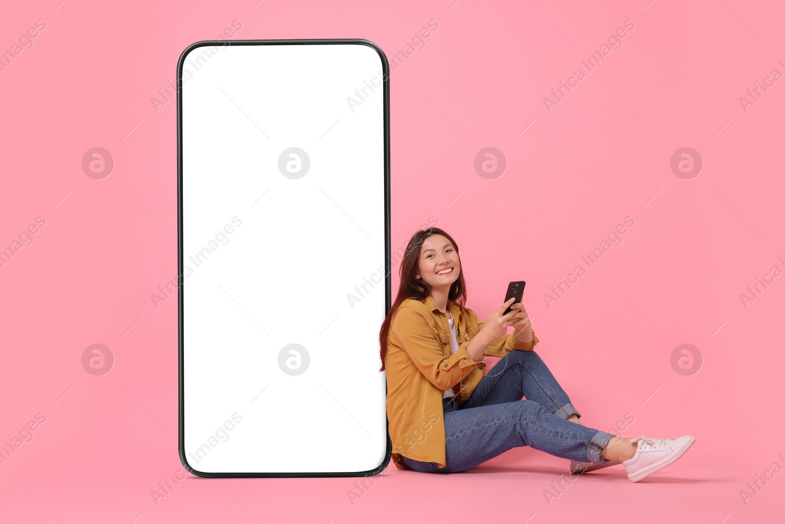 Image of Happy smiling woman with smartphone sitting near big mobile phone with blank screen on pink background. Mockup for design