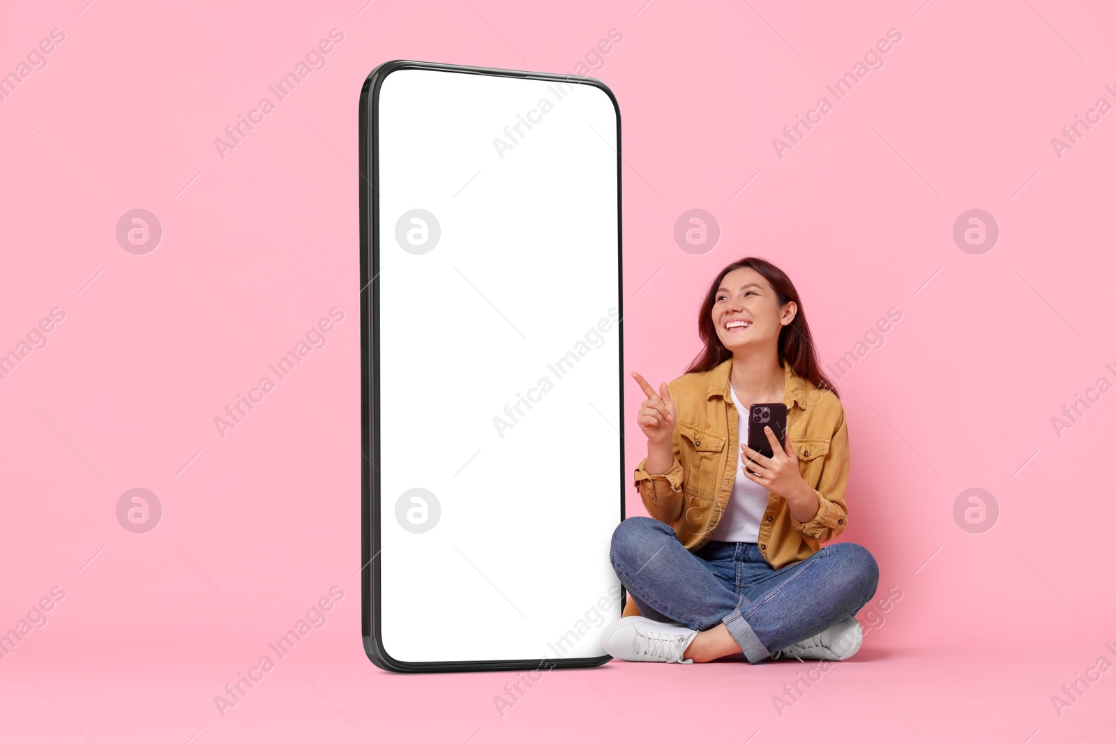 Image of Happy smiling woman with smartphone pointing at big mobile phone with blank screen on pink background. Mockup for design