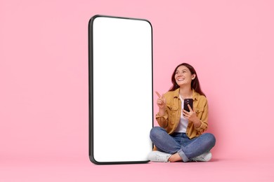 Happy smiling woman with smartphone pointing at big mobile phone with blank screen on pink background. Mockup for design