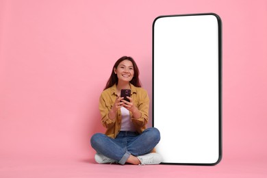 Image of Happy smiling woman with smartphone sitting near big mobile phone with blank screen on pink background. Mockup for design