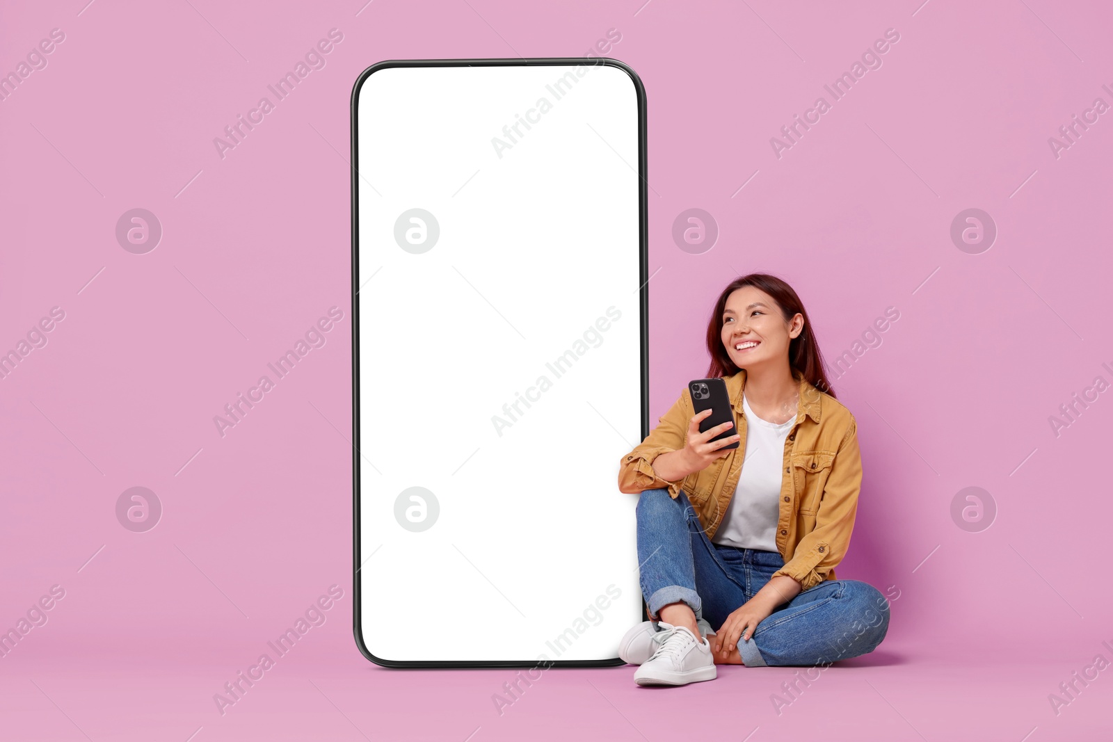 Image of Happy smiling woman with smartphone sitting near big mobile phone with blank screen on lilac background. Mockup for design