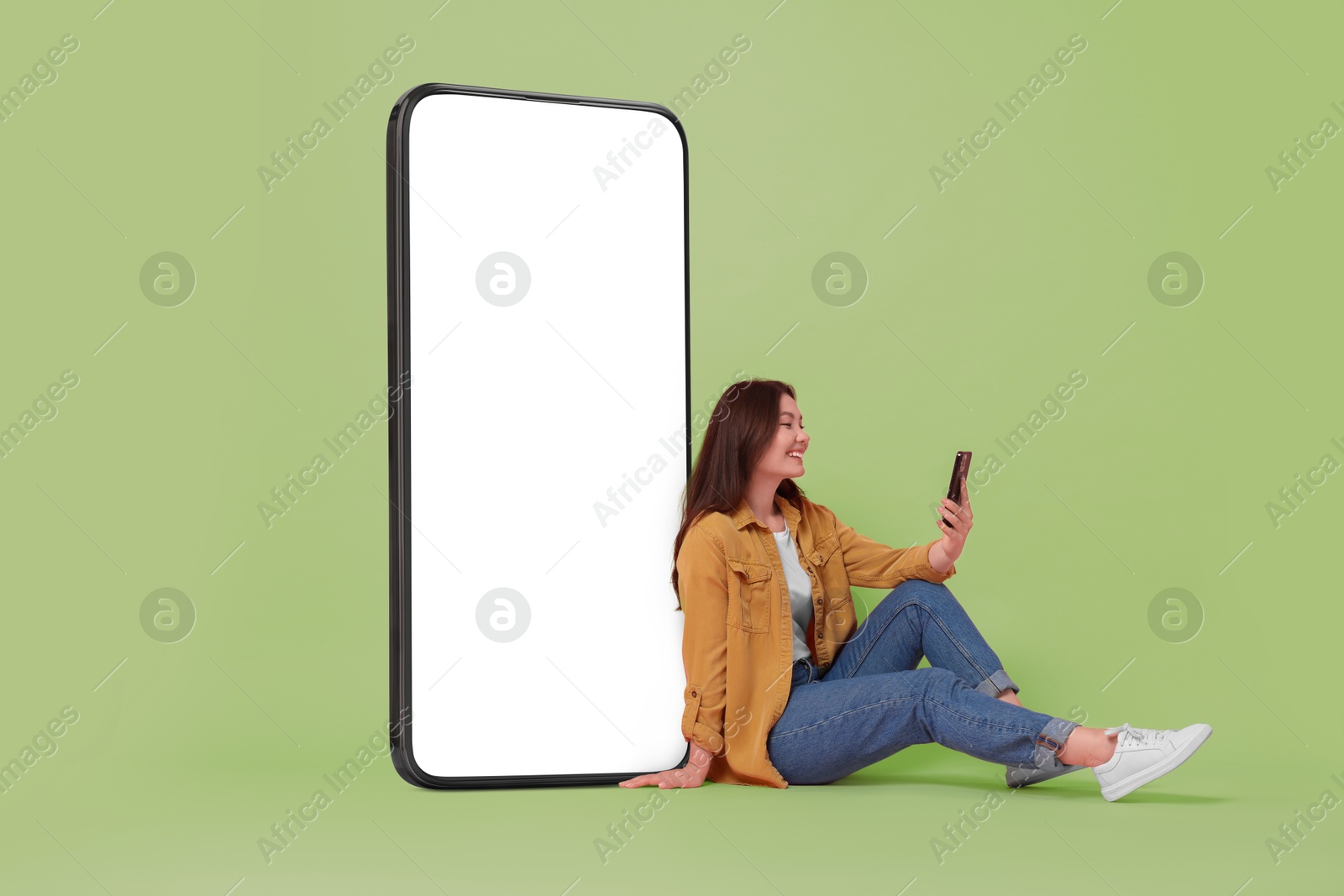 Image of Happy smiling woman using smartphone while sitting near big mobile phone with blank screen on light green background. Mockup for design