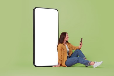 Happy smiling woman using smartphone while sitting near big mobile phone with blank screen on light green background. Mockup for design