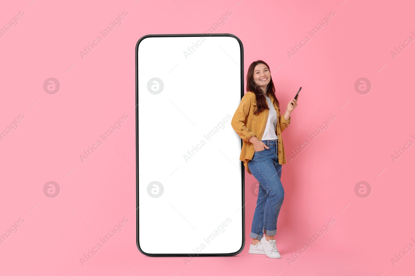 Image of Happy smiling woman with smartphone standing near big mobile phone with blank screen on pink background. Mockup for design