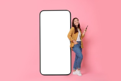 Image of Happy smiling woman with smartphone standing near big mobile phone with blank screen on pink background. Mockup for design