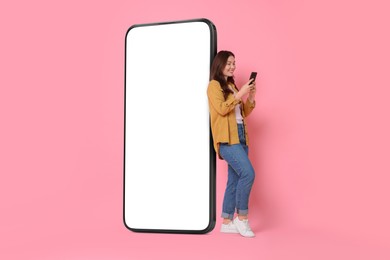 Image of Happy smiling woman using smartphone while standing near big mobile phone with blank screen on pink background. Mockup for design