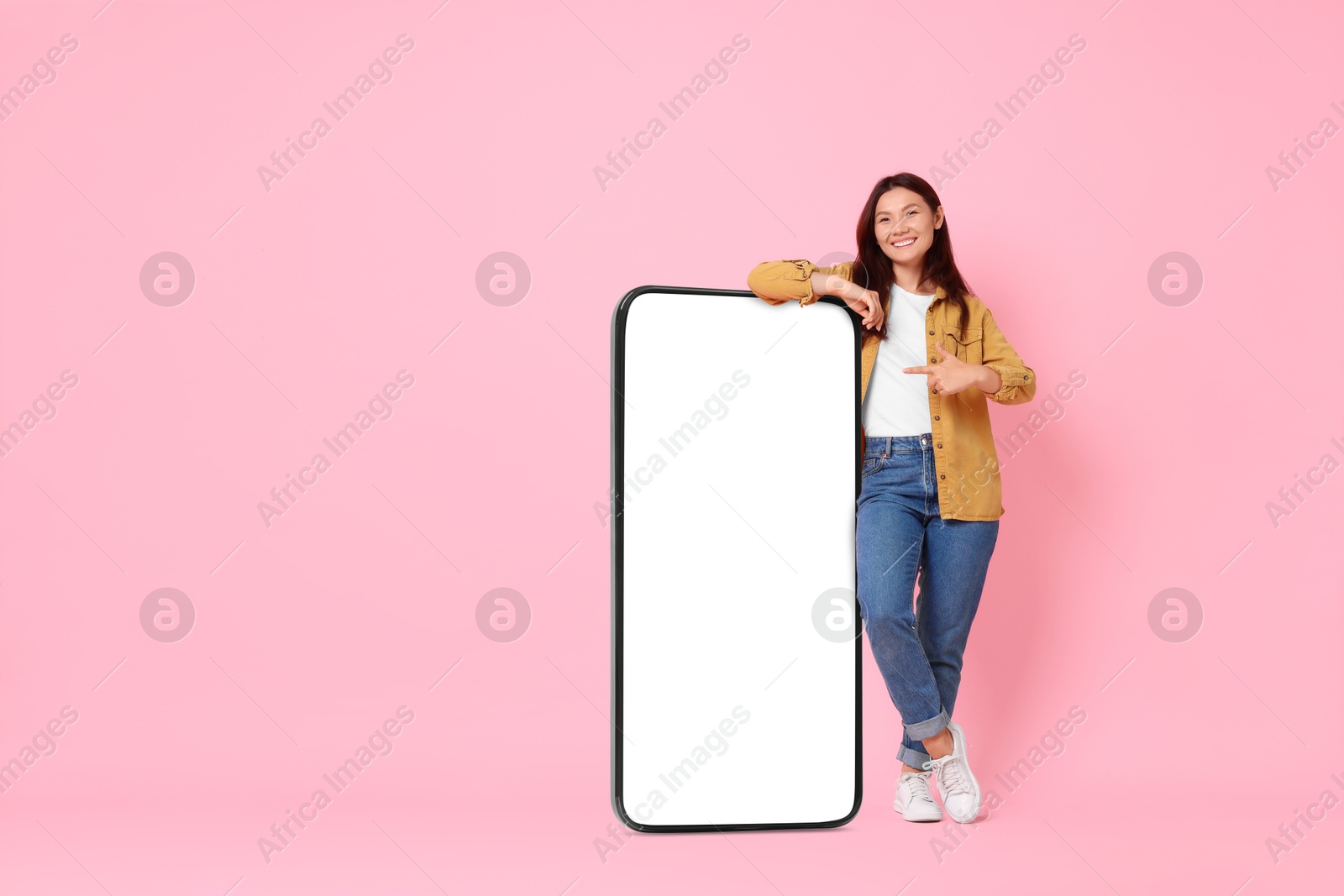 Image of Happy smiling woman pointing at big mobile phone with blank screen on pink background. Mockup for design