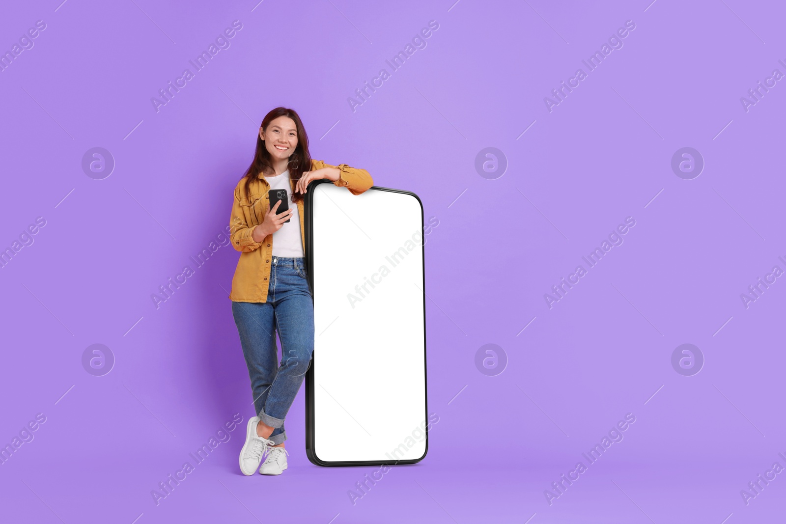 Image of Happy smiling woman with smartphone standing near big mobile phone with blank screen on violet background. Mockup for design