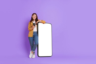 Image of Happy smiling woman with smartphone standing near big mobile phone with blank screen on violet background. Mockup for design