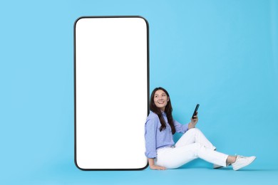 Happy smiling woman with smartphone sitting near big mobile phone with blank screen on light blue background. Mockup for design