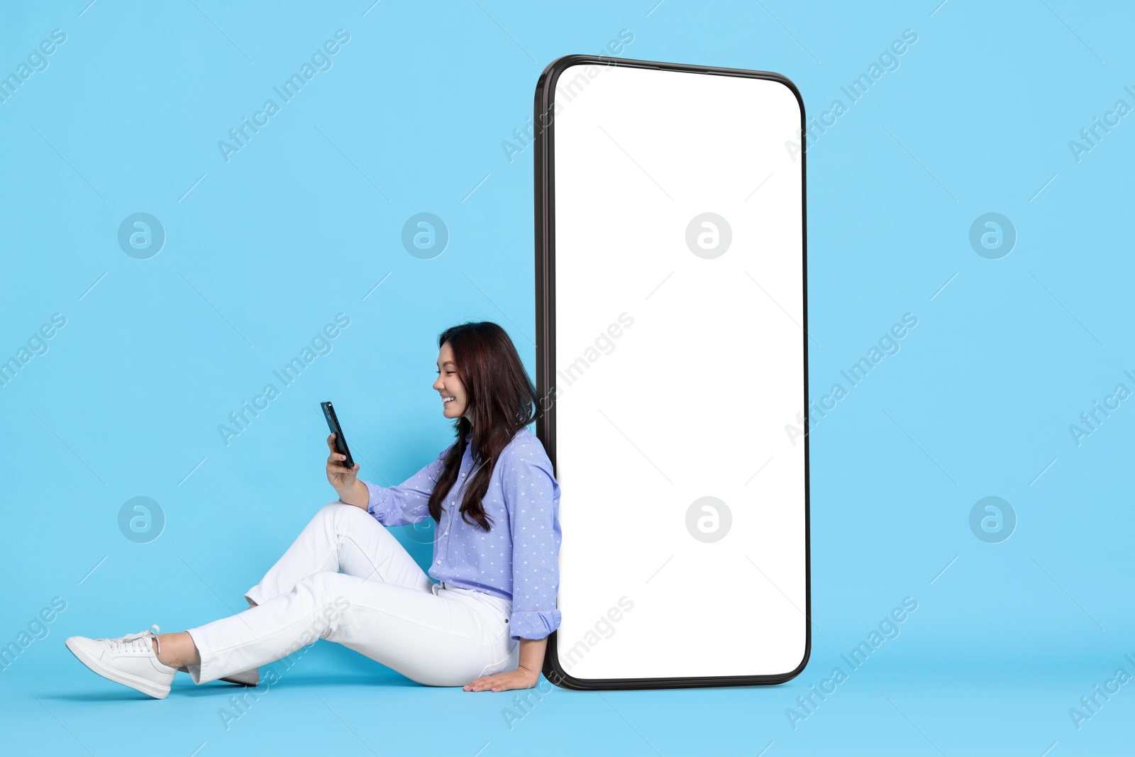 Image of Happy smiling woman using smartphone while sitting near big mobile phone with blank screen on light blue background. Mockup for design