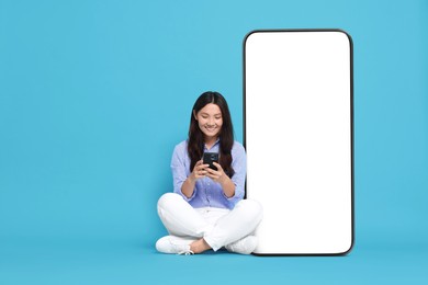 Image of Happy smiling woman using smartphone while sitting near big mobile phone with blank screen on light blue background. Mockup for design