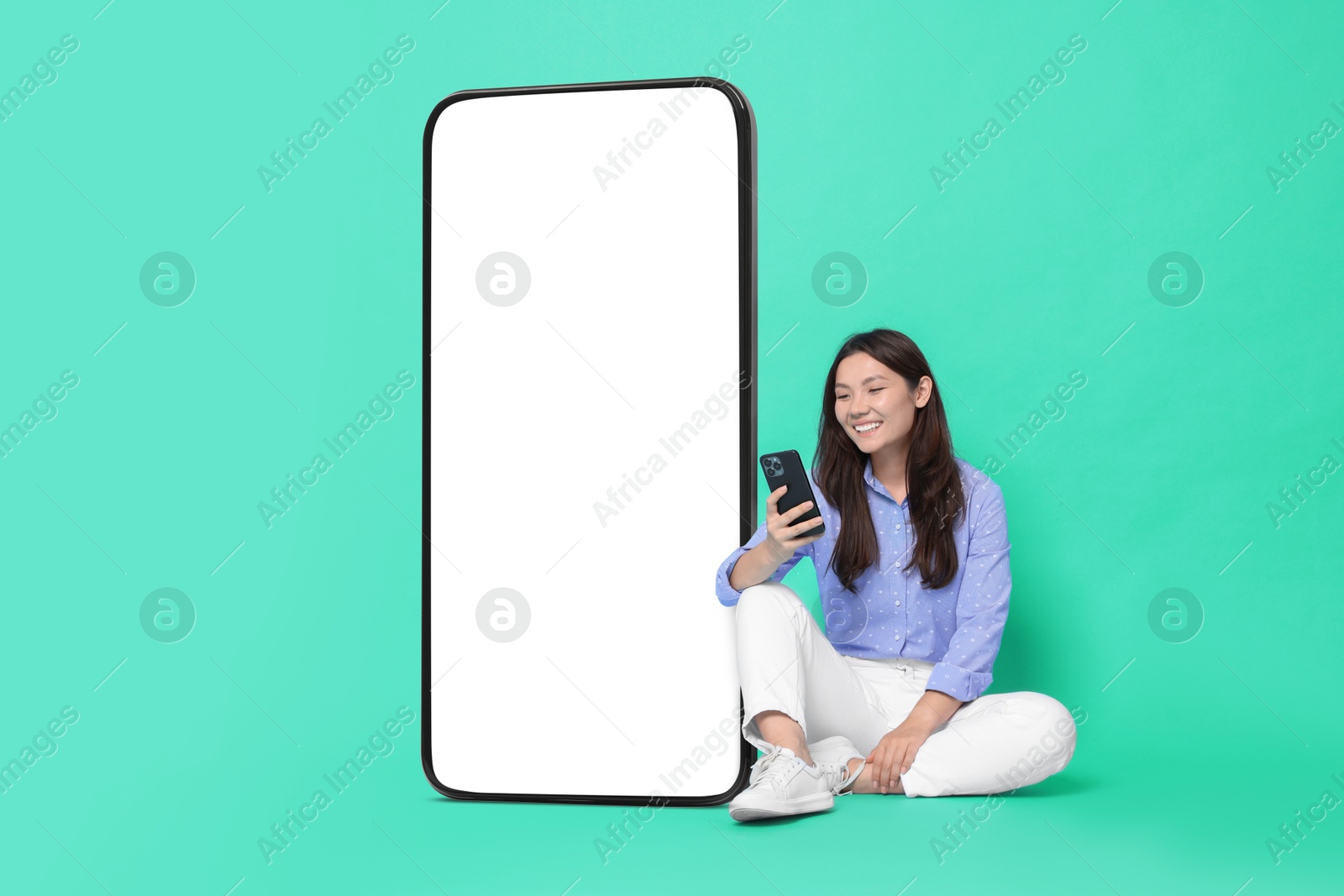 Image of Happy smiling woman using smartphone while sitting near big mobile phone with blank screen on turquoise background. Mockup for design