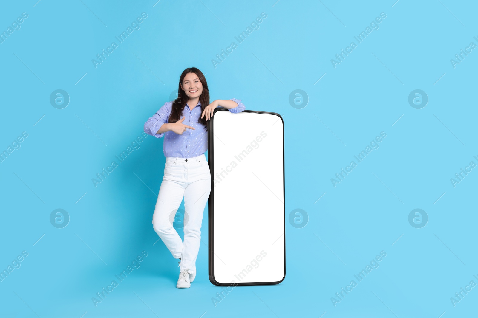 Image of Happy smiling woman pointing at big mobile phone with blank screen on light blue background. Mockup for design