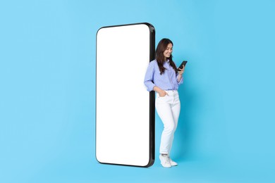 Image of Happy smiling woman using smartphone while standing near big mobile phone with blank screen on light blue background. Mockup for design