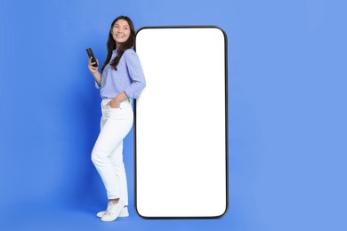 Image of Happy smiling woman with smartphone standing near big mobile phone with blank screen on blue background. Mockup for design