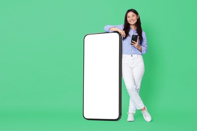 Image of Happy smiling woman with smartphone standing near big mobile phone with blank screen on aquamarine background. Mockup for design