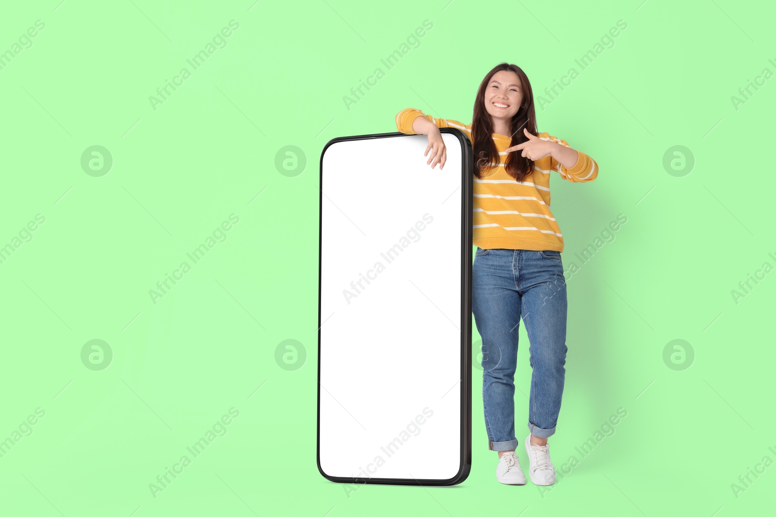 Image of Happy smiling woman pointing at big mobile phone with blank screen on light green background. Mockup for design