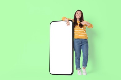 Image of Happy smiling woman pointing at big mobile phone with blank screen on light green background. Mockup for design