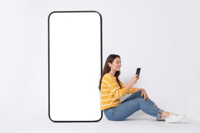 Happy smiling woman using smartphone while sitting near big mobile phone with blank screen on while background. Mockup for design