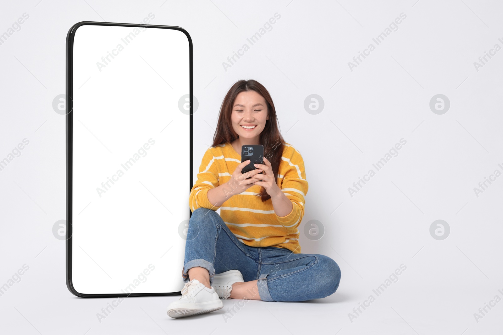 Image of Happy smiling woman using smartphone while sitting near big mobile phone with blank screen on white background. Space for design