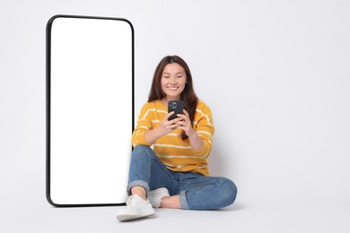 Happy smiling woman using smartphone while sitting near big mobile phone with blank screen on white background. Space for design